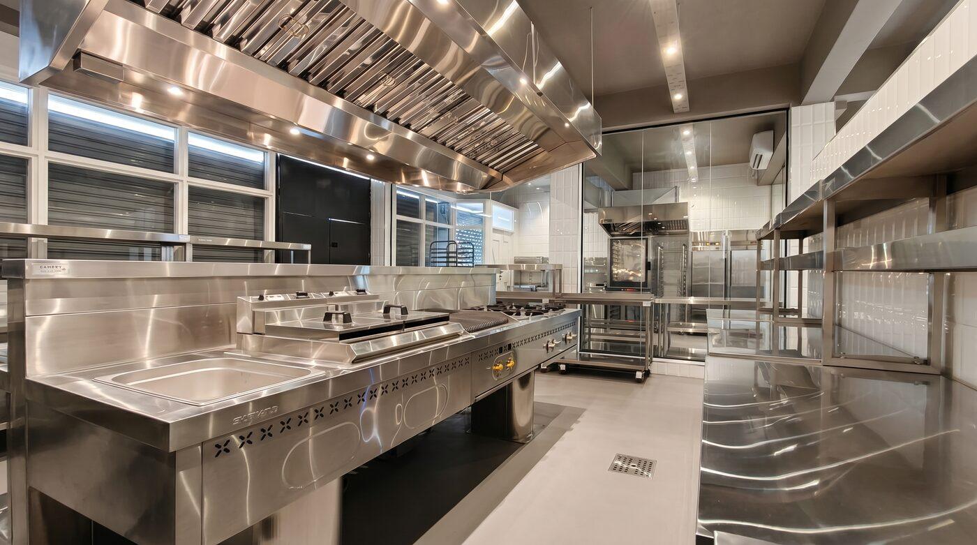 Inside the kitchen: stainless steel prep counters, ranges, and extraction hoods under bright light.